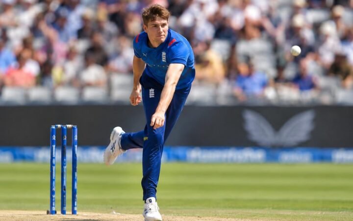 Jogador de críquete inglês Will Jacks em ação durante arremesso, vestindo uniforme azul, com o campo e espectadores desfocados ao fundo.