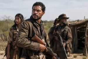Cena da série "Cangaço Novo" com três pessoas em destaque segurando armas, vestindo roupas simples e sujas, em um ambiente rural de caatinga com uma construção rústica ao fundo.
